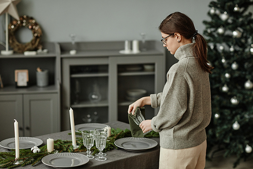 Side view portrait of young woman setting up plates on dining table decorated for Christmas with fir branches and candles in grey tones, copy space