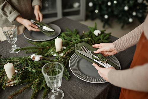 High angle close up of young woman setting up plates on dining table decorated for Christmas with fir branches and candles in grey tones, copy space