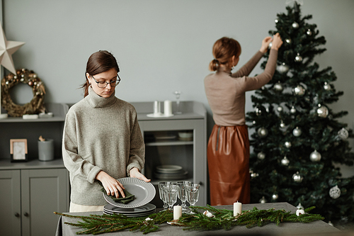 Portrait of young woman setting up plates on dining table decorated for Christmas with fir branches and candles in grey tones, copy space