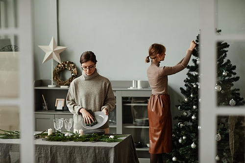 Portrait of two young women setting up dining room decorated for Christmas with fir branches and candles in grey tones, copy space