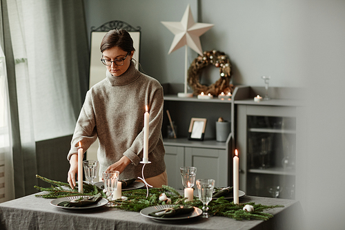 Portrait of young woman setting up dining table decorated for Christmas with fir branches and candles in grey tones, copy space