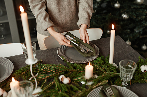 Closeup of unrecognizable woman setting up dining table for Christmas decorated with fir branches and candles in grey tones, copy space