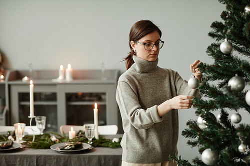 Waist up portrait of young woman decorating Christmas tree at home in elegant silver and grey tones, copy space