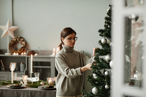 Waist up portrait of young woman hanging ornaments on Christmas tree at home in elegant silver and grey tones, copy space