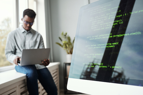 Blurred portrait of young African-American man using laptop while sitting by window in software development office, code screen in foreground, copy space