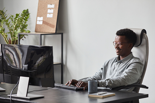 Portrait of smiling African-American man using computer while sitting at desk in office, software developer concept, copy space