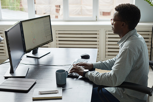 Side view portrait of focused African-American man using computer while sitting at desk in office, software developer concept, copy space