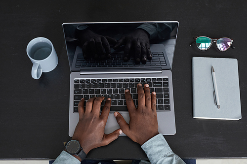 Minimal top view of unrecognizable African-American man using laptop while working at black desk background, focus on hands at keyboard, copy space