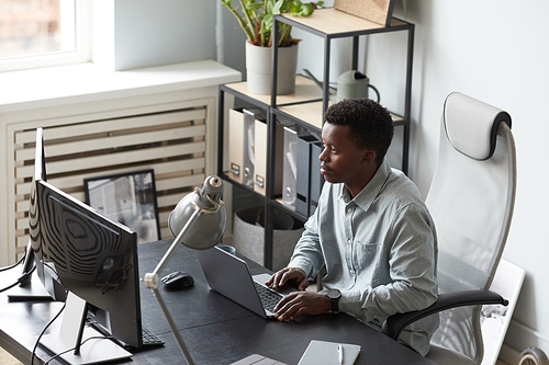 High angle portrait of focused African-American man using computer with two screens while sitting at desk in office, software developer concept, copy space