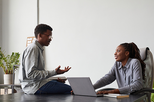 Minimal side view portrait of two African-American young people chatting in office while discussing work project by computers, copy space