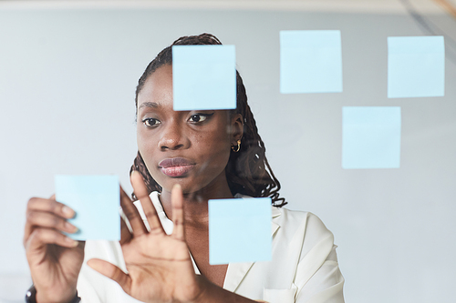 Minimal portrait of young African-American businesswoman putting sticker notes on glass wall while planning project in office, copy space