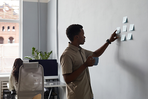 Side view portrait of young African-American man placing sticker notes on wall while planning project in office, copy space