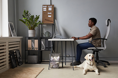 Full length portrait of young African-American man working in office with dog laying on floor, pet friendly workspace, copy space