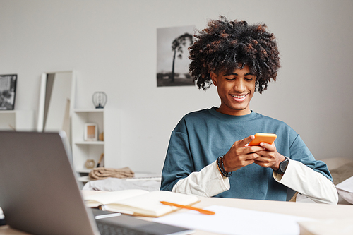 Portrait of African-American teenage boy using smartphone and smiling while studying at home or in college dorm, copy space