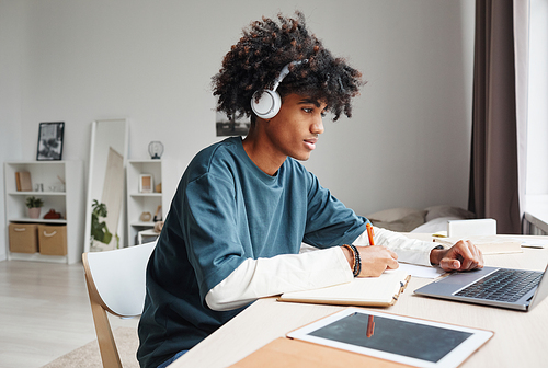 Side view portrait of African-American teenage boy studying at home or in college dorm and using laptop, copy space