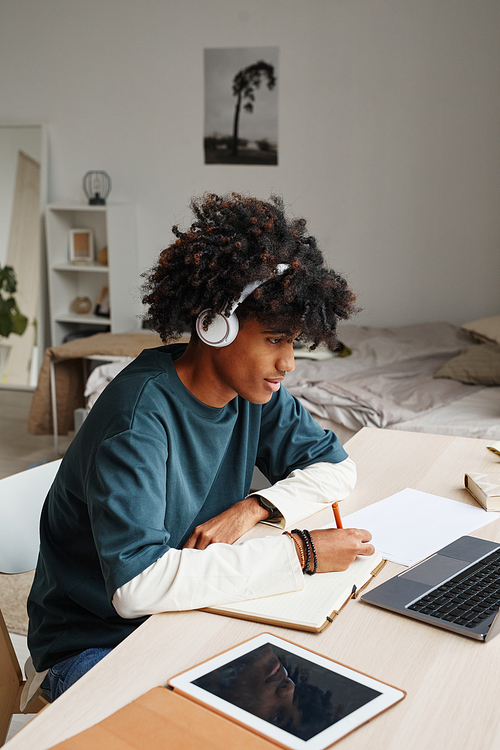 Vertical portrait of African-American teenage boy studying at home or in college dorm and using laptop