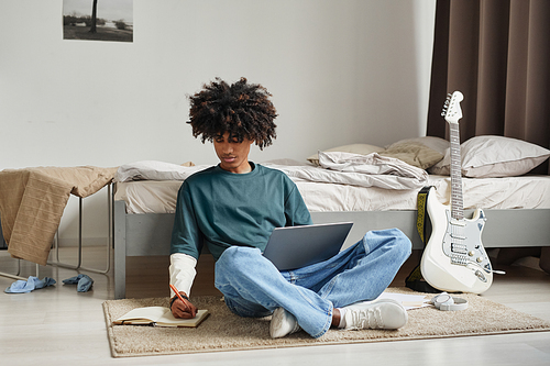 Full length portrait of teenage African-American student sitting on floor at home or in college dorm and studying , copy space