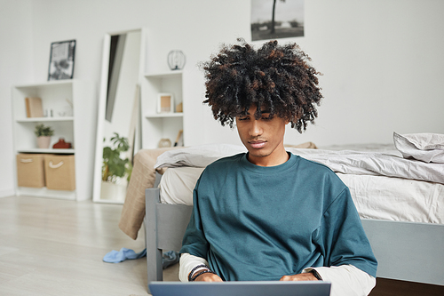 Portrait of teenage African-American student sitting on floor at home or in college dorm and using laptop, copy space