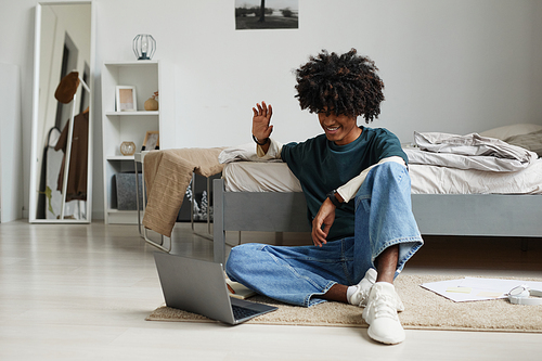 Portrait of teenage African-American boy sitting on floor at home or in college dorm and waving to camera wile speaking by video chat online, copy space