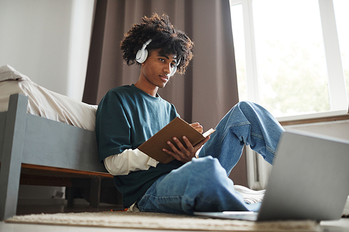 Low angle portrait of teenage African-American student sitting on floor at home or in college dorm and studying , copy space