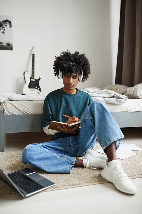 Vertical full length portrait of teenage African-American student sitting on floor at home or in college dorm and studying