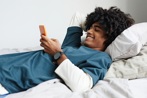 Side view portrait of young African-American man lying on bed at home and smiling while using smartphone