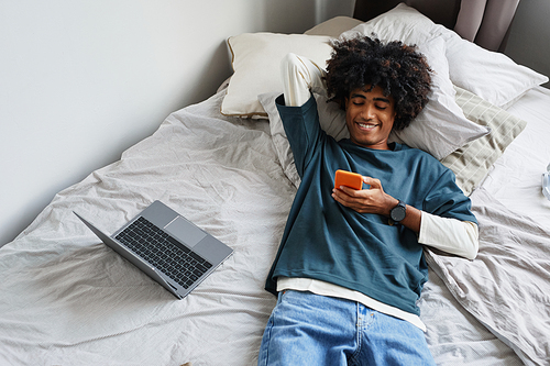 Above view portrait of young African-American man lying on bed at home and smiling while using smartphone, copy space