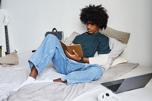 Full length portrait of young African-American man sitting on bed at home and writing in notebook, copy space