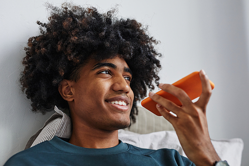 Close up of young African-American man speaking by smartphone on bed and smiling, focus on natural hair, copy space