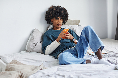 Minimal full length portrait of young African-American man using smartphone on bed at home, copy space