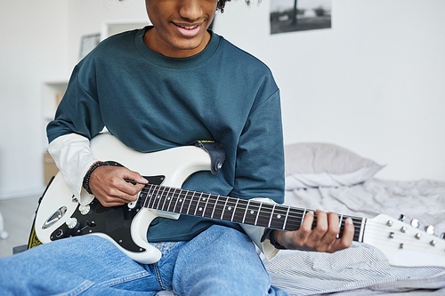 Cropped portrait of smiling African-American teenager playing guitar while sitting on bed at home, copy space