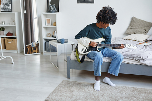 Full length portrait of smiling African-American teenager playing guitar while sitting on bed at home, copy space