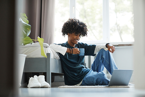 Full length portrait of smiling African-American teenager using video chat while sitting on floor at home, copy space