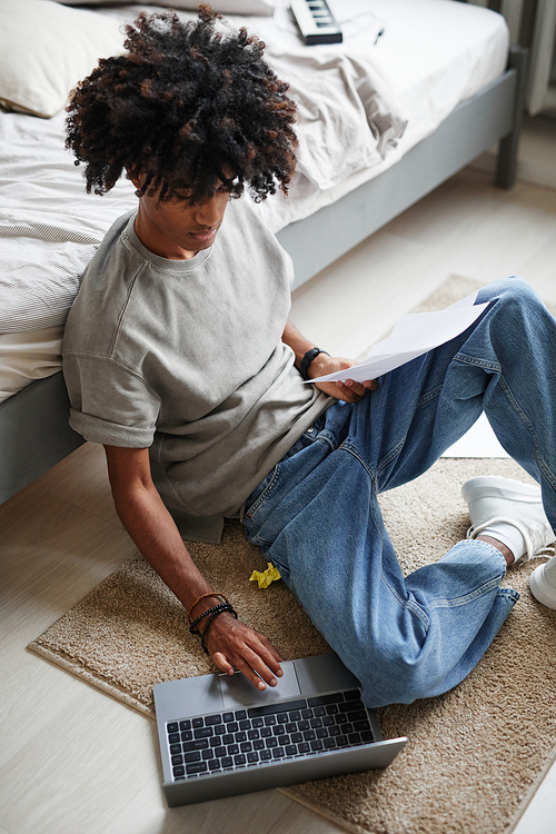 Vertical portrait of African-American teenager using laptop while sitting on floor at home