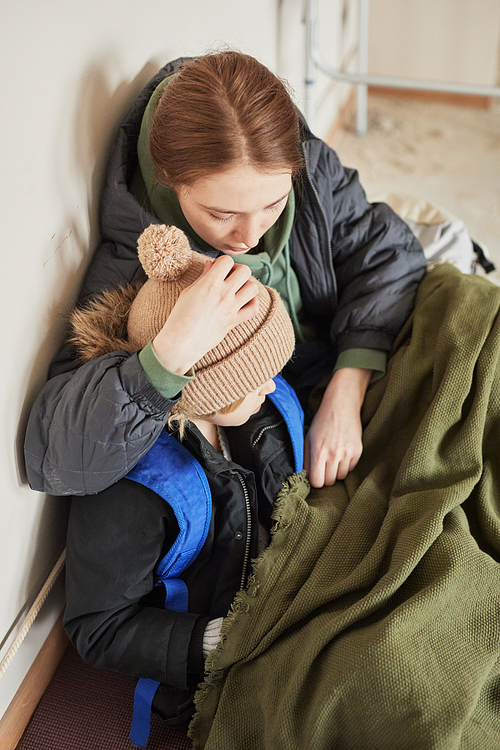 Portrait of young mother embracing son while hiding together in refugee shelter and trying to keep warm