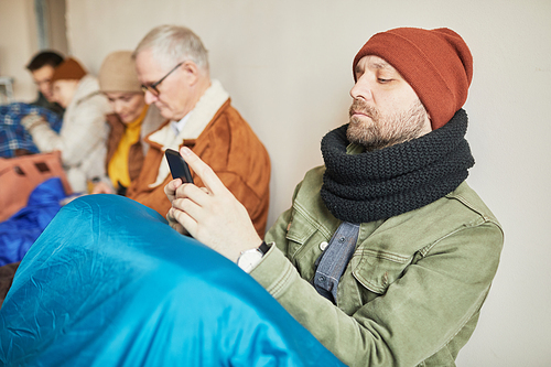 Side view portrait of bearded Caucasian man hiding in refugee shelter and holding smartphone, copy space