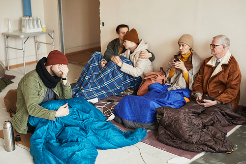Group of distressed Caucasian refugees hiding in shelter during war or crisis covered with blankets on floor