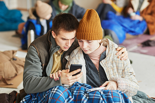Front view portrait of young Caucasian couple hiding in refugee shelter covered with blanket on floor and holding smartphone