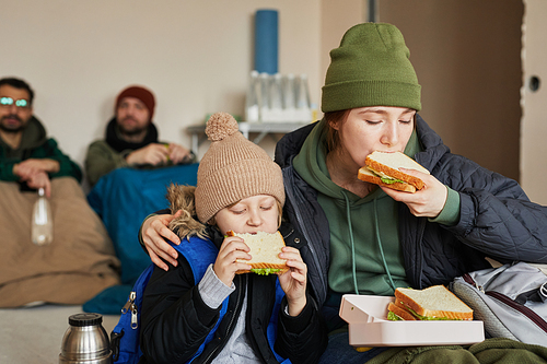 Portrait of Caucasian young mother and son eating sandwiches in refugee shelter during war or crisis, copy space