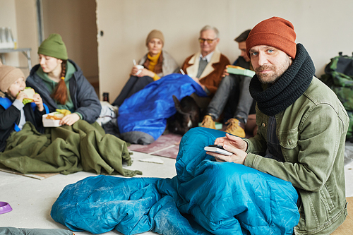 Portrait of Caucasian man hiding in refugee shelter and looking at camera while sitting on floor covered with blanket, copy space