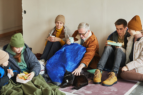 Group of Caucasian refugees eating food while hiding in shelter on floor covered with blankets