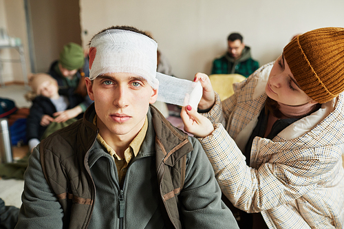 Front view portrait of Caucasian wounded man in refugee shelter with young woman bandaging head