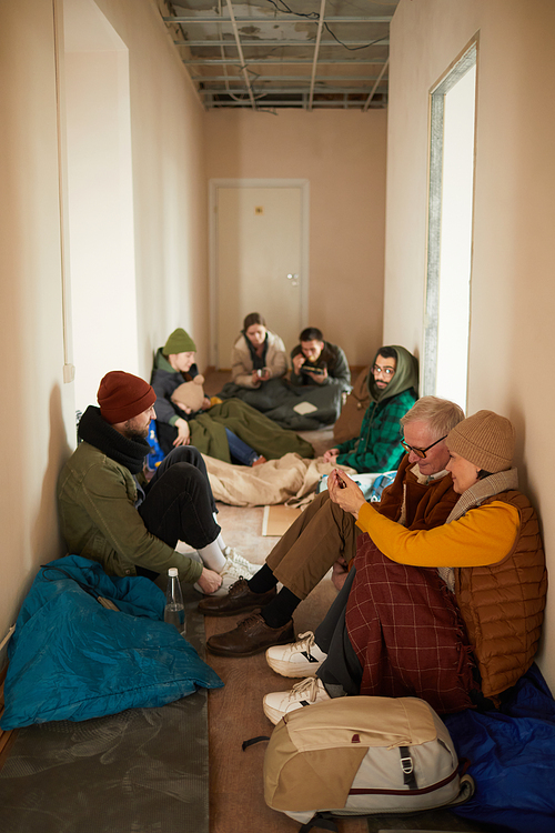 Vertical shot of hallway in refuge shelter filled with group of people sitting on floor by sleeping bags