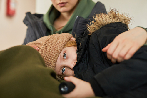 Portrait of blonde Caucasian child lying in mothers lap at refugee shelter and looking at camera with blue eyes