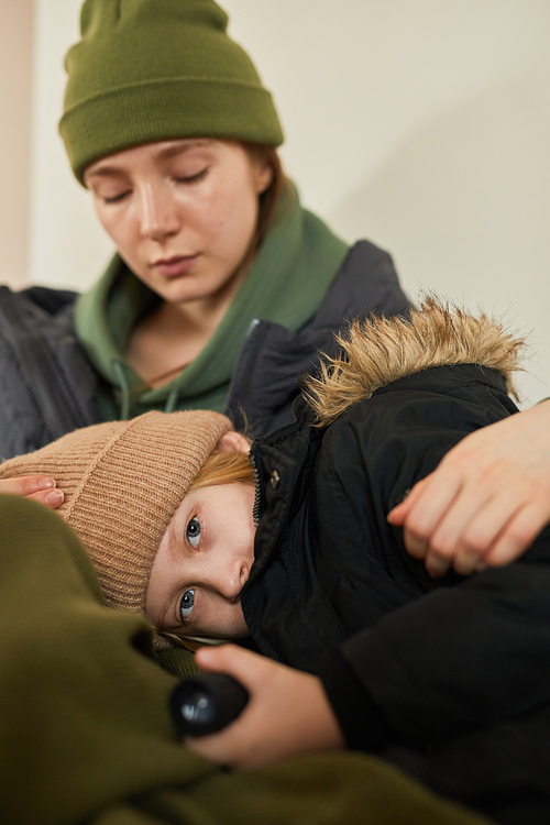 Vertical portrait of blonde Caucasian child lying in mother's lap at refugee shelter and looking at camera