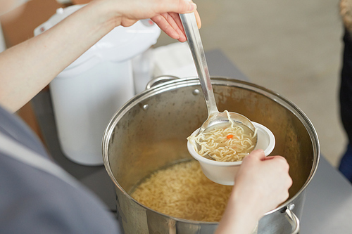 Close up of young woman pouring soup into bowl while volunteering at help center for people in need, copy space