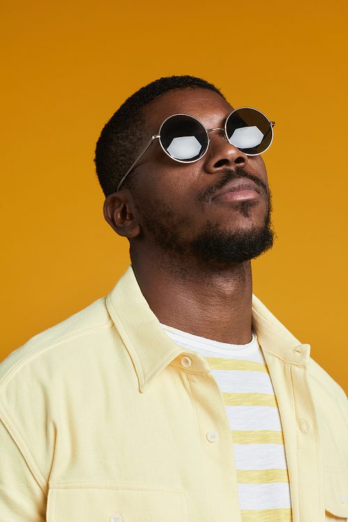 Vertical portrait of handsome African-American man wearing sunglasses while posing against yellow background in studio