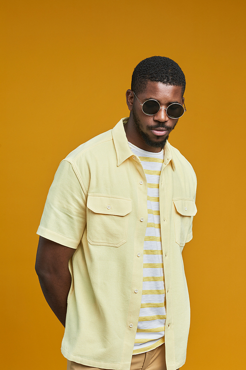 Vertical waist up portrait of handsome African-American man wearing sunglasses while posing against yellow background in studio