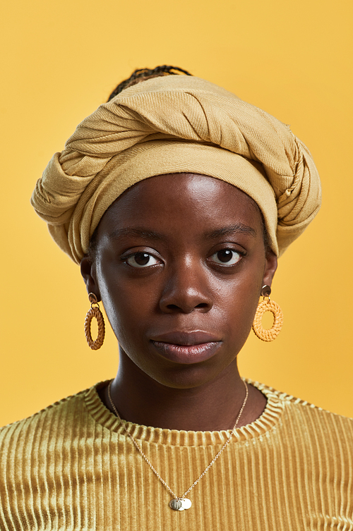 Vertical close up portrait of young African-American woman wearing head wrap and ethnic jewelry looking at camera while posing against yellow background in studio