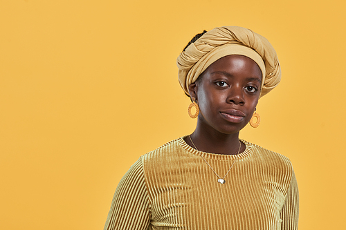 Minimal portrait of young African-American woman wearing head wrap and looking at camera while posing against yellow background in studio, copy space